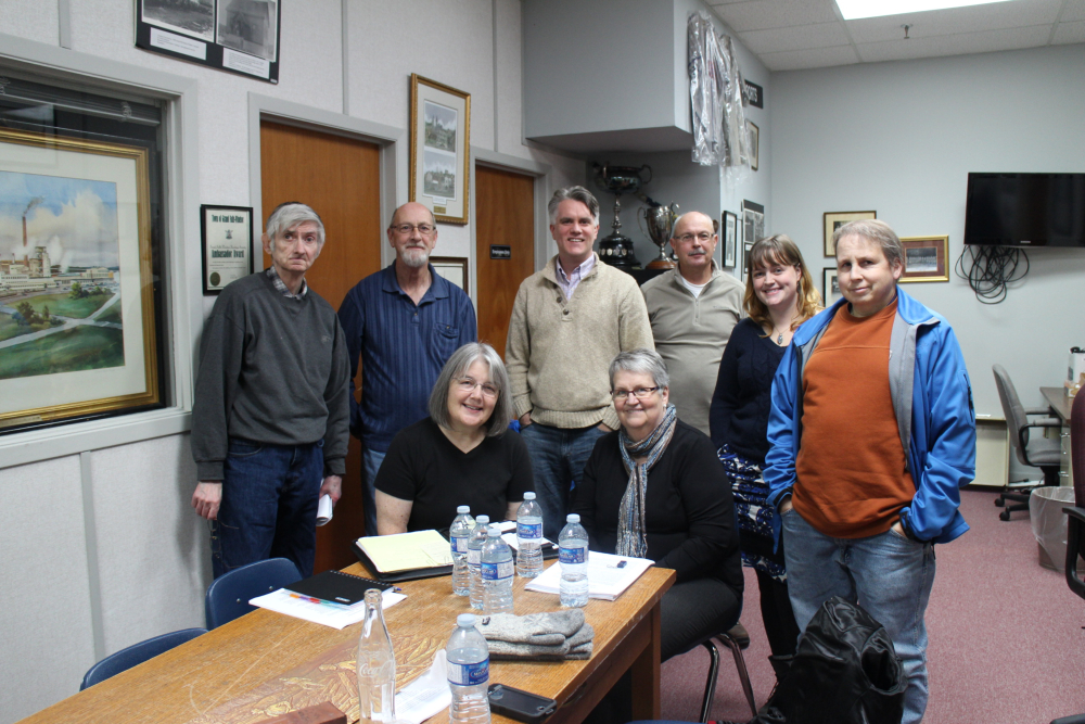 Colour photograph. Standing from left to right are four men, one woman, and another man. Sitting at table are two women.