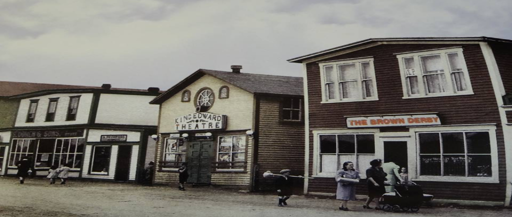 Colour archival photograph. Brown Derby, King Edward Theatre, A. Peckford, S. Cohen & Sons, and Purity Cafe. Kirk Pomeroy is carrying a birch broom, Daisy Bennett is wearing a blue-grey coat, and Mrs. Pomeroy is pushing a stroller.