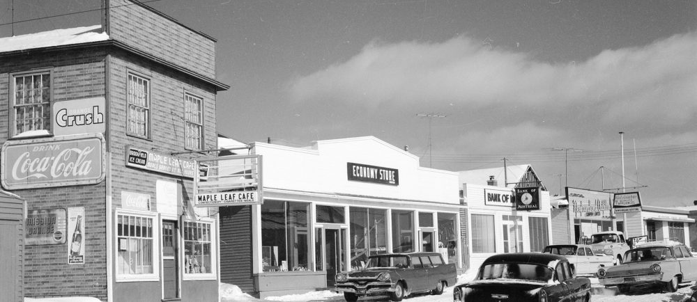 Black and white archival photograph. Street view. Maple Leaf Cafe, Economy Store, Bank of Montreal, Newfoundland Outfitting, and Windsor Post Office visible. Several cars are in the streets and there is snow on the ground.