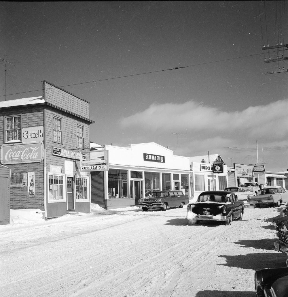 Black and white archival photograph. Street view. Maple Leaf Cafe, Economy Store, Bank of Montreal, Newfoundland Outfitting, and Windsor Post Office visible. Several cars are in the streets and there is snow on the ground.