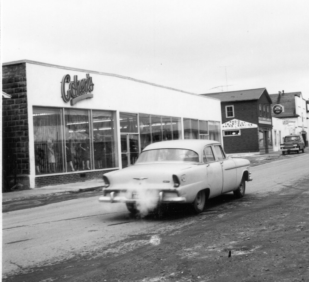 Black and white archival photograph. Street view. Cohen’s, Main Street looking east towards Great Eastern Oil, and E. Becker’s shop. Women's manikins visible in the windows on the left side of Cohen’s main doors. Light car in the centre of the photo.