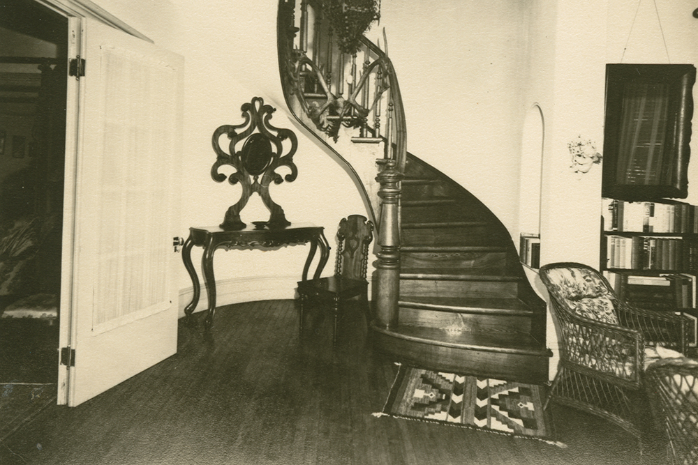 Back-and-white photograph of the entrance hall of Manoir Fraser with a wooden staircase in the centre.