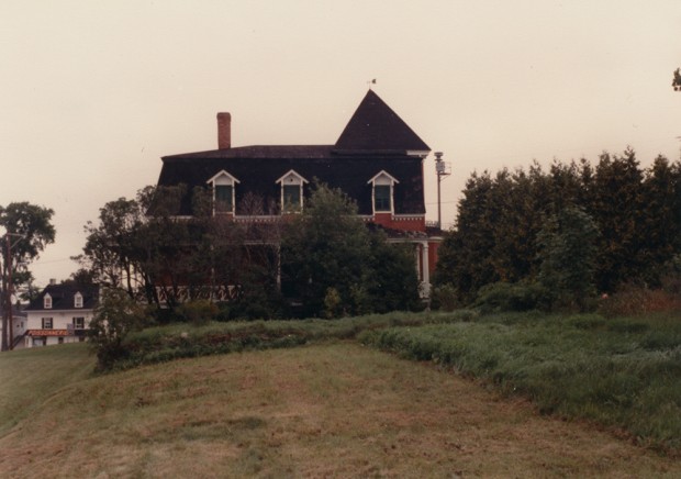 Colour photograph. In the centre, an empty house. In the foreground, an overgrown yard.