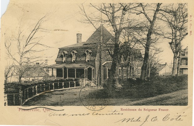 A black-and-white postcard showing a large elegant house with a veranda, bay window, and mansard roof. A few mature trees line the drive to the house.