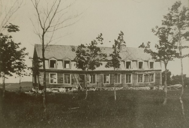 Black-and-white photograph of a long house, apparently empty. The mansard roof has many dormer windows.