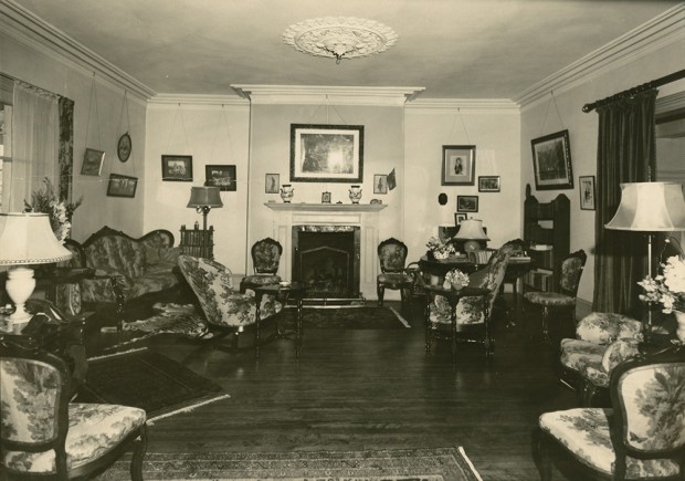 Black-and-white photograph of a crowded living room. The furniture is arranged along the walls on the right and left, and a fireplace is in the wall at the rear.