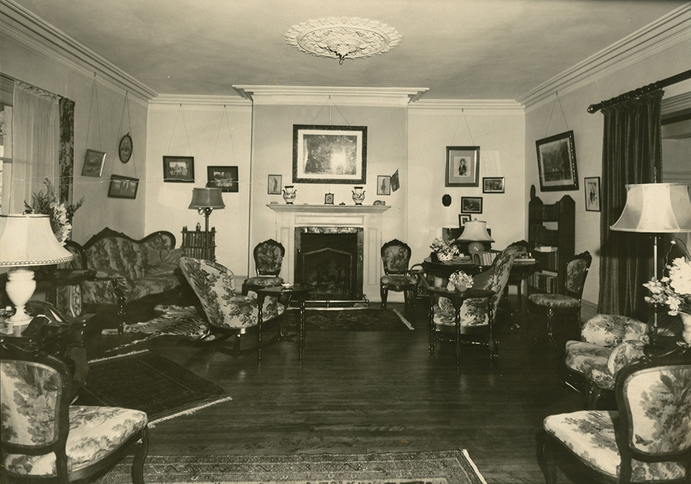 Black-and-white photograph of a crowded living room. The furniture is arranged along the walls on the right and left, and a fireplace is in the wall at the rear.