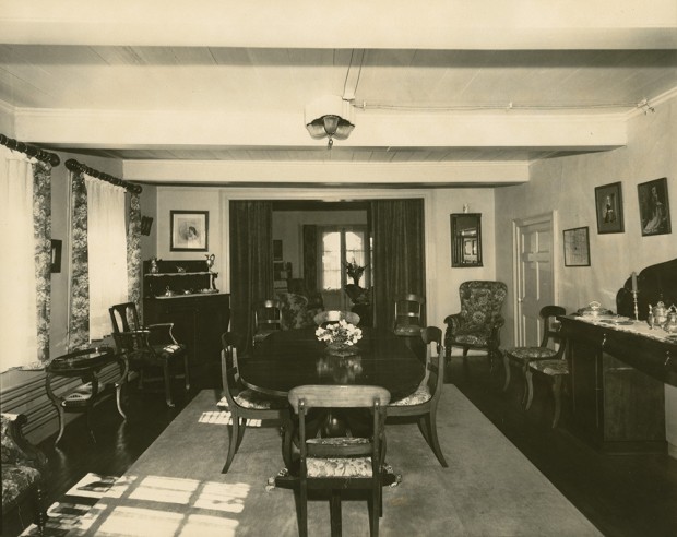 Black-and-white photograph of the dining room. The table is in the centre, on a large carpet. A long sideboard is against the wall to the right. The drawing room is connected to this room.