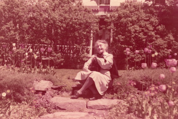 Colour photograph of an elderly woman sitting on steps in a flower garden.