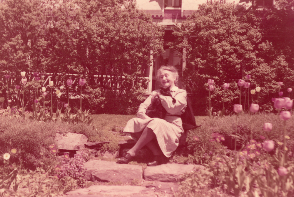 Colour photograph of an elderly woman sitting on steps in a flower garden.