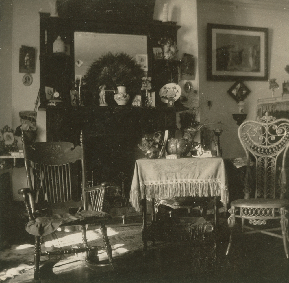 Black-and-white photograph of a drawing room. Two chairs and a table in the foreground are in front of a fireplace with a large mirror over it. There are numerous pictures and decorative objects on the wall and the mantelpiece.