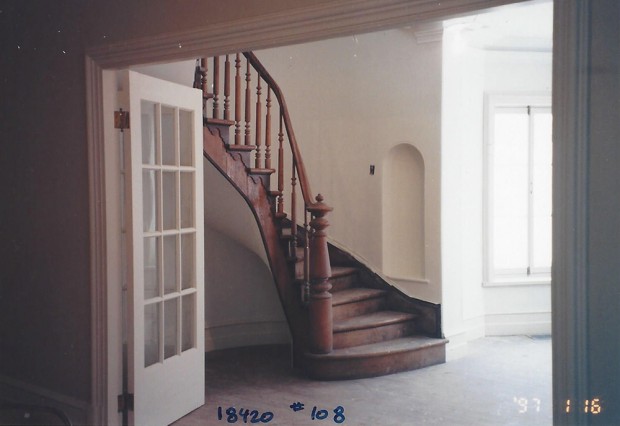 Colour photograph of the empty entrance hall of Manoir Fraser with a wooden staircase in the centre.