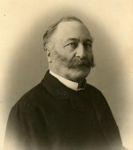 Black-and-white head-and-shoulders photograph of a man with a huge moustache and greying hair, dressed in a black suit.