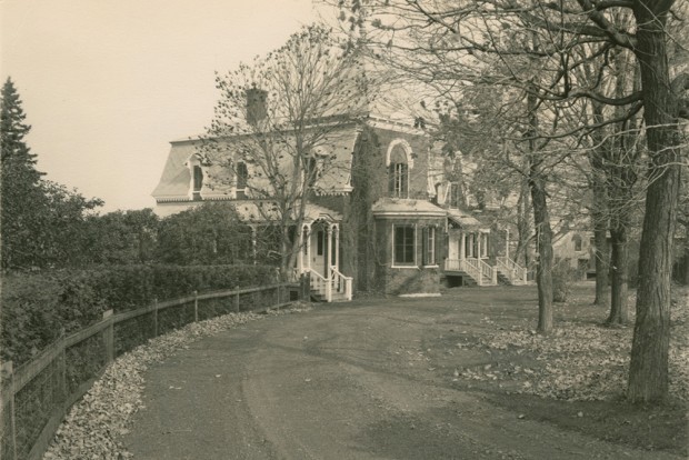 Black-and-white photograph. A road in the foreground leads to a house surrounded by trees.