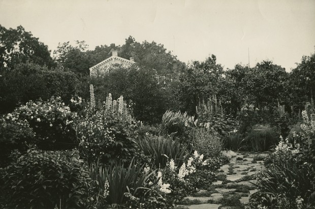 Black-and-white photograph of a lush flowering garden with a flagstone walkway in the foreground. Many mature trees hide the house in the background.