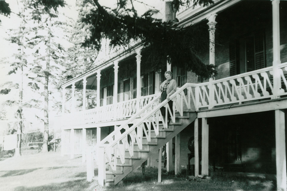 Black-and-white photograph of an elderly woman standing on the staircase leading to the large veranda that wraps around Manoir Fraser.
