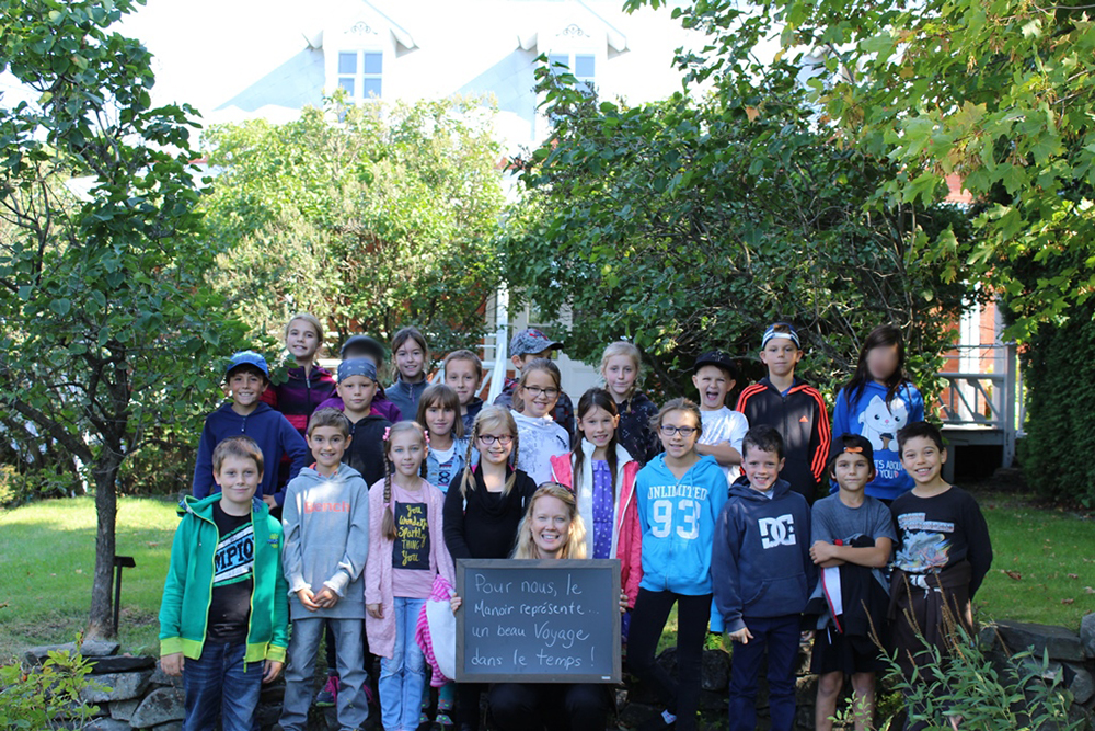 Colour photograph of a group of some 20 schoolchildren standing among trees with a house in the background. Their teacher, in front, holds a blackboard with the words For us, the Manoir represents a beautiful voyage in time!