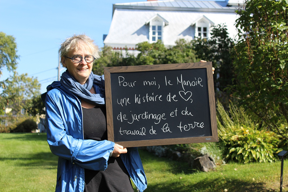 Colour photograph of a woman holding a small backboard with the words To me, the Manoir represents an affair of the heart, a story of gardening and working the soil. A large house with a mansard roof is in the background.