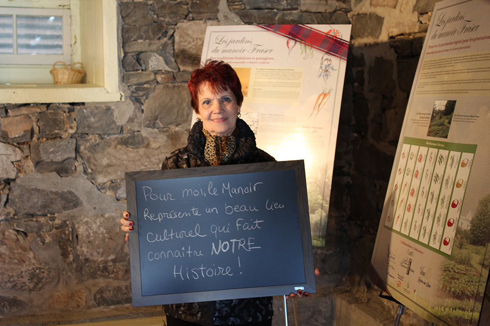 Colour photograph of a woman standing inside the manor holding a small blackboard with the words To me, the Manoir represents a lovely cultural site that promotes knowledge of OUR history.