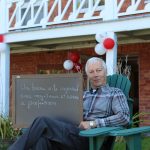 Colour photograph of a man sitting in a garden chair, holding a small blackboard on which he has written what the Manoir represents to him: A lovely, restful place, with a profusion of plants and birds. A red brick house is in the background.