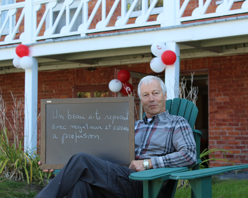 Colour photograph of a man sitting in a garden chair, holding a small blackboard on which he has written what the Manoir represents to him: A lovely, restful place, with a profusion of plants and birds. A red brick house is in the background.