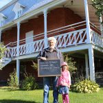 Colour photograph of a man holding a small blackboard with the words: To me, the Manoir represents memories of youth. A little girl stands beside him, and a large brick house with a big veranda is behind him.