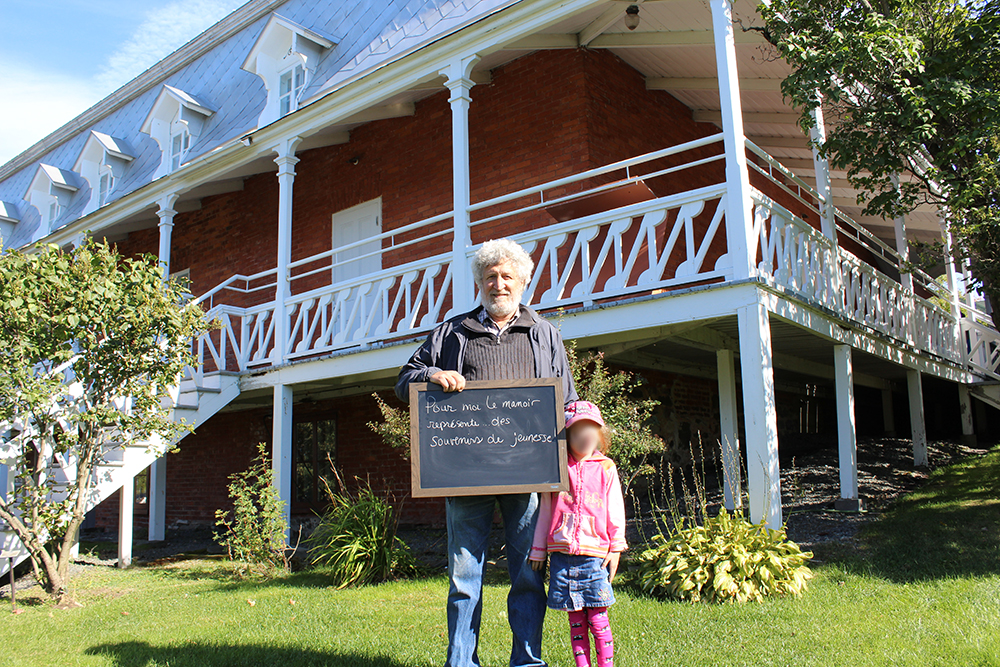 Colour photograph of a man holding a small blackboard with the words: To me, the Manoir represents memories of youth. A little girl stands beside him, and a large brick house with a big veranda is behind him.