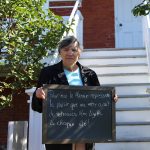 Colour photograph of a woman holding a small blackboard with the words To me, the Manoir represents the pleasure my mother used to have meeting Madame Lizotte every summer. A stairway leading to a veranda is in the background.