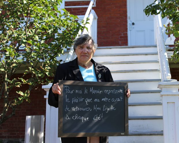 Colour photograph of a woman holding a small blackboard with the words To me, the Manoir represents the pleasure my mother used to have meeting Madame Lizotte every summer. A stairway leading to a veranda is in the background.