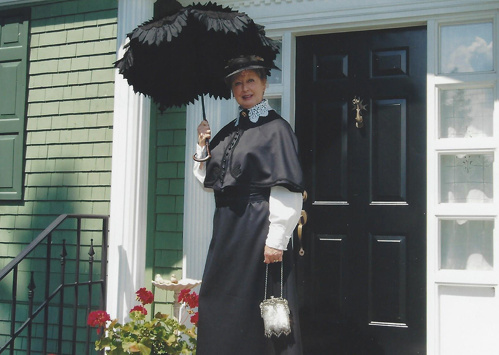 Colour photograph of a woman in a Victorian dress in front of a house painted green.