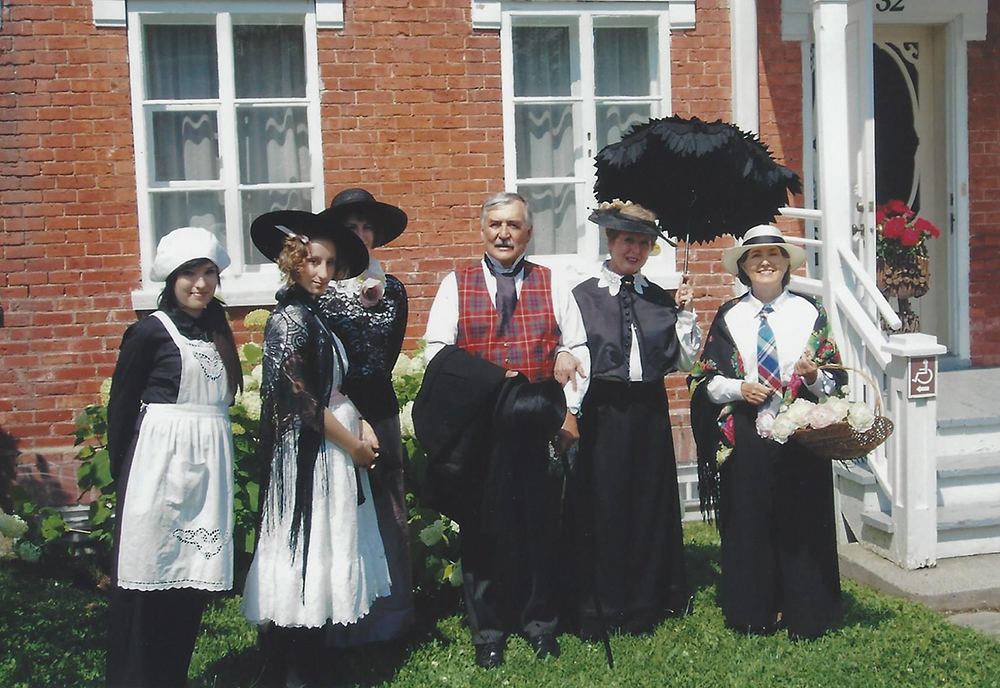 Colour photograph of one man and five women dressed in period costumes, posing for the camera in front of a brick house.