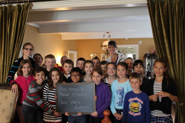 Colour photograph of a group of about 20 smiling schoolchildren standing between the manor drawing room and the dining room. One holds a small blackboard with the words To us, the Manoir is a part of our history.