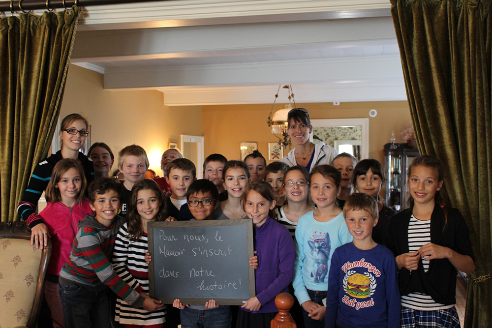 Colour photograph of a group of about 20 smiling schoolchildren standing between the manor drawing room and the dining room. One holds a small blackboard with the words To us, the Manoir is a part of our history.