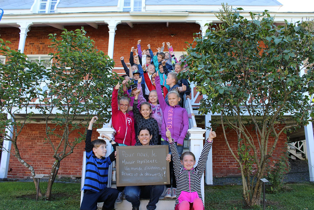 Colour photograph of about 20 smiling schoolchildren with their arms raised, on a staircase leading to a red brick house. Their teacher is holding a small blackboard with the words To us, the Manoir represents a world of stories, treasures, and discoveries!