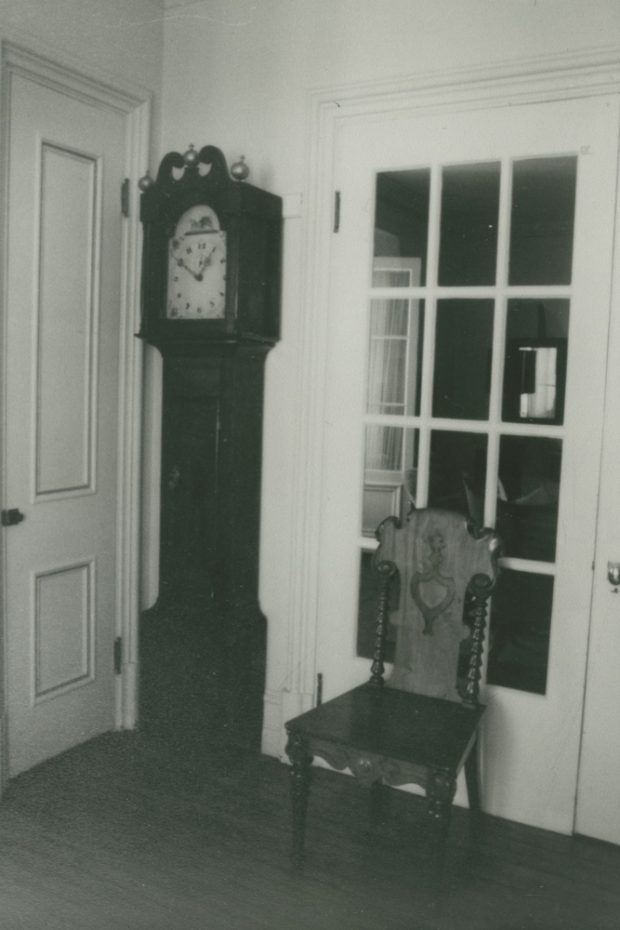 Black-and-white photograph. A grandfather clock stands in a corner. Beside it is a chair, placed against a glass door.