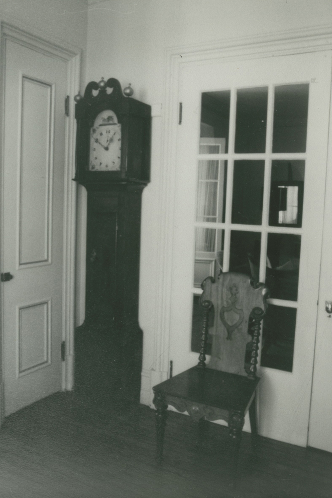 Photo noir et blanc. Une horloge adossée contre un mur et une chaise adossée contre une porte vitrée.