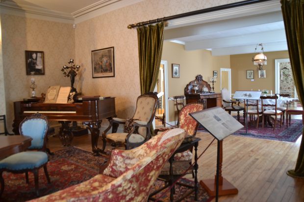 Colour photograph of the drawing room, decorated in the Victorian style. A baby grand piano is in the corner. An opening in the wall gives onto the dining room. An explanatory panel is on a stand on the right.