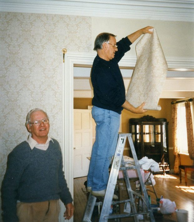 Colour photograph of two men, one on a step-ladder, putting up wallpaper in the drawing room.