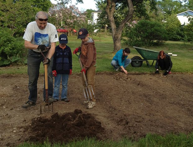 A man in the foreground shows two boys how to turn over the soil for the vegetable garden. In the background, a woman and a young girl are picking up rocks from the garden.