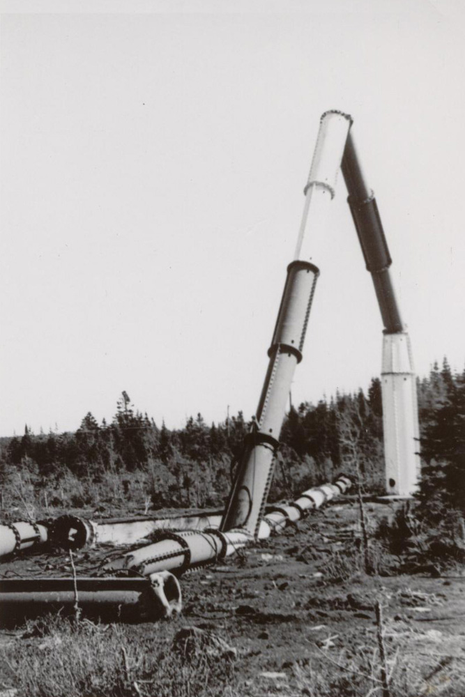 Sections of round metal tubing, some standing connected to a base, some lying on the ground, with fir trees in the background.