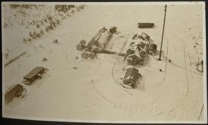 An aerial view of three buildings and an antenna encircled by fencing, with additional outbuildings beyond. Snow covers the ground.