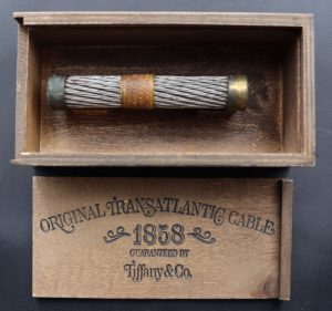 Petit morceau de câble métallique tressé dans une boîte en bois. Le couvercle de la boîte porte la mention « Câble transatlantique original. 1858. Garanti par Tiffany & Co. » (traduction).