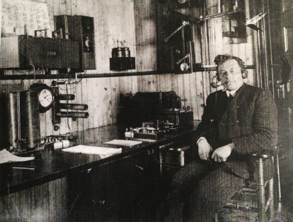 A man in a suit sits facing the camera beside a desk topped by wireless equipment and neat piles of paper. He wears a set of wired headphones and smokes a pipe.