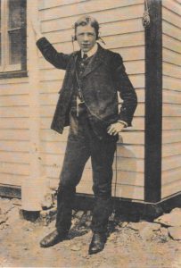 A young man in a suit and vest stands before a clapboard building, his right arm raised to clasp a flagpole. He wears headphones and looks directly into the camera.