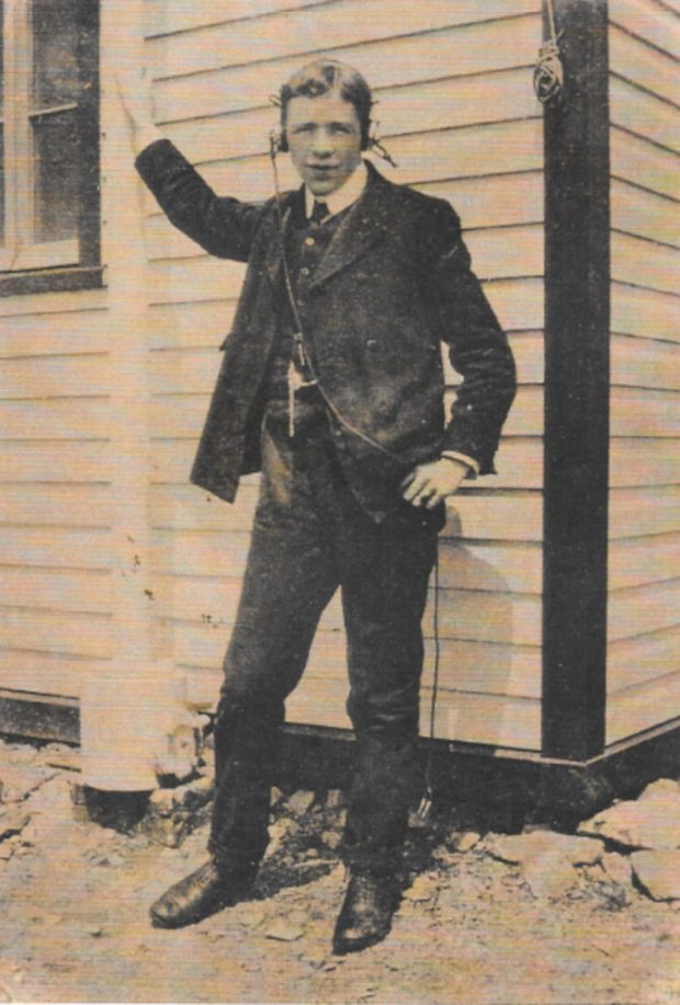 A young man in a suit and vest stands before a clapboard building, his right arm raised to clasp a flagpole. He wears headphones and looks directly into the camera.