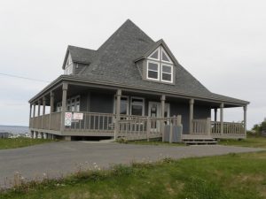 Maison grise moderne avec des lucarnes et une galerie tout autour de la maison, située sur un terrain dégagé en bordure de l’océan.