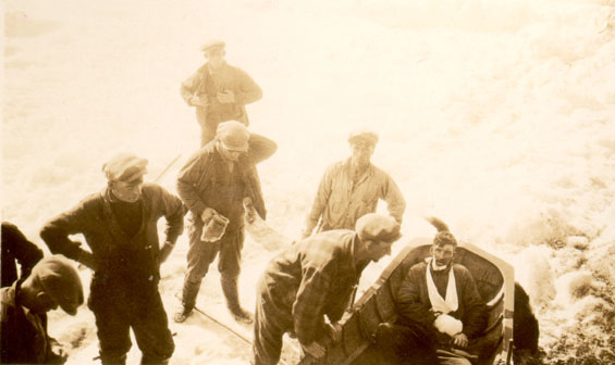 Seven men, dressed in caps, boots, and coats, stand on an ice field. One leans over a small boat with an injured man in it. The hurt man’s right hand and jaw are bandaged.