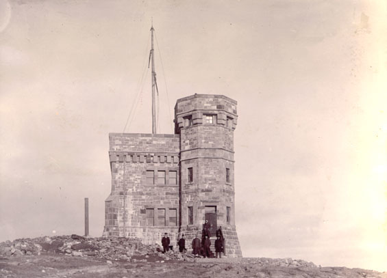 A group of people stand in front of a two-storey stone building on a barren rocky hilltop. The building has a taller turret on the right and a mast on the rooftop at left.