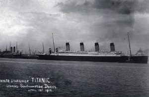 A large ship with four funnels steams to the right out of a port, leaving behind smaller vessels. Written on the photograph is: “White Star liner ‘Titanic.’ Leaving Southampton Docks, April 10th, 1912.”
