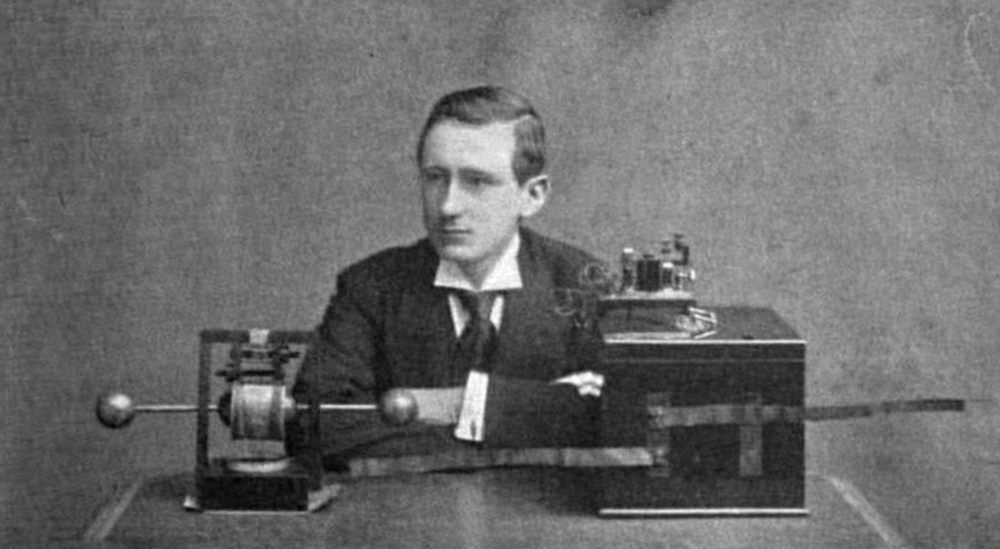 A young man sits with his crossed arms resting on a table. Radio equipment sits on the table in front of him.